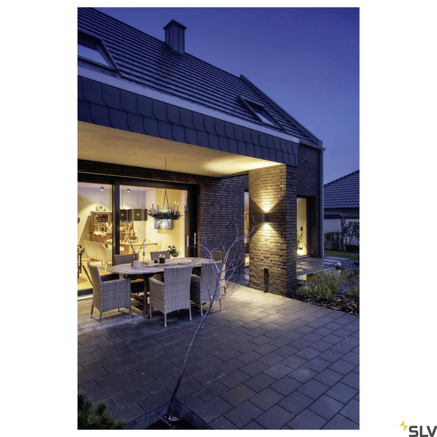 Ein modernes Haus bei Dämmerung mit großer Fensterfront und Terrasse, auf der ein Esstisch mit Stühlen steht.