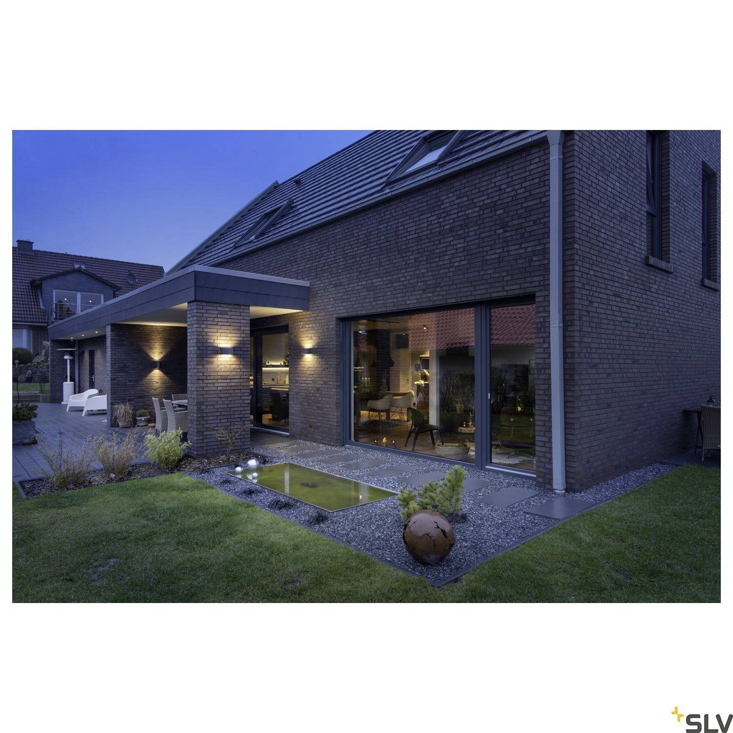 Ein modernes Einfamilienhaus bei Dämmerung, beleuchtet mit Außenleuchten. Der Garten hat einen kleinen Teich und gepflegten Rasen.