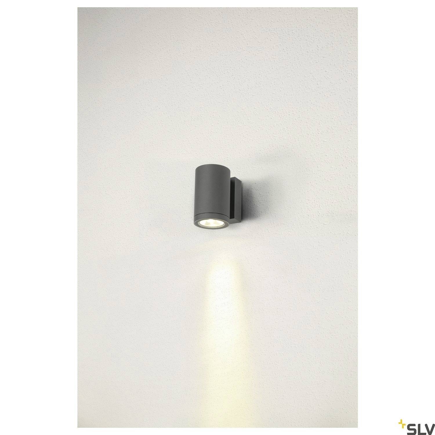 Wandleuchte mit zylindrischem Gehäuse, strahlt warmes Licht nach unten auf eine weiße Wand. Geeignet für moderne Innenräume.