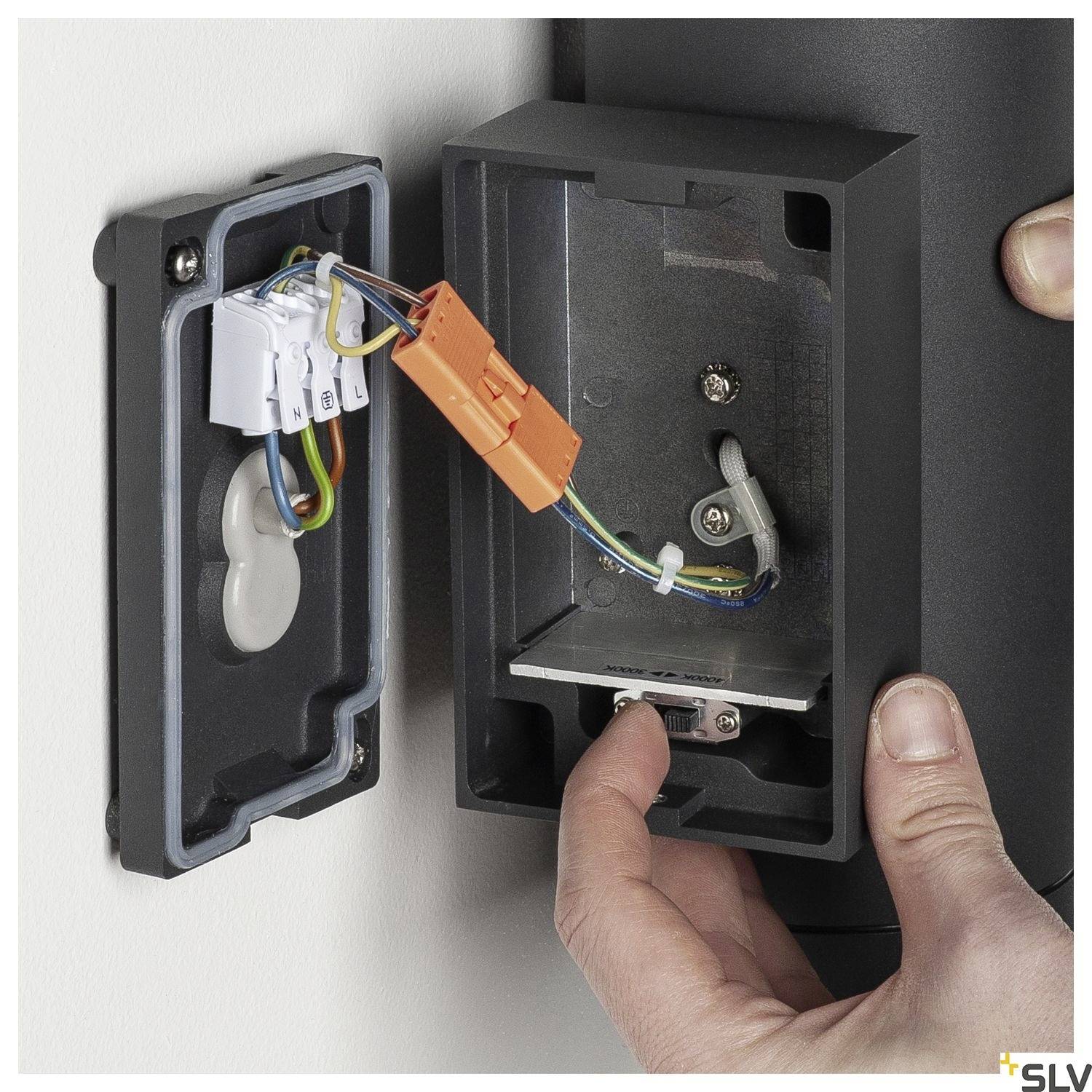 Jemand montiert eine Wandlampe. Die Rückseite ist geöffnet und zeigt bunte Drähte, die an ein elektrisches Anschlussstück angeschlossen sind.