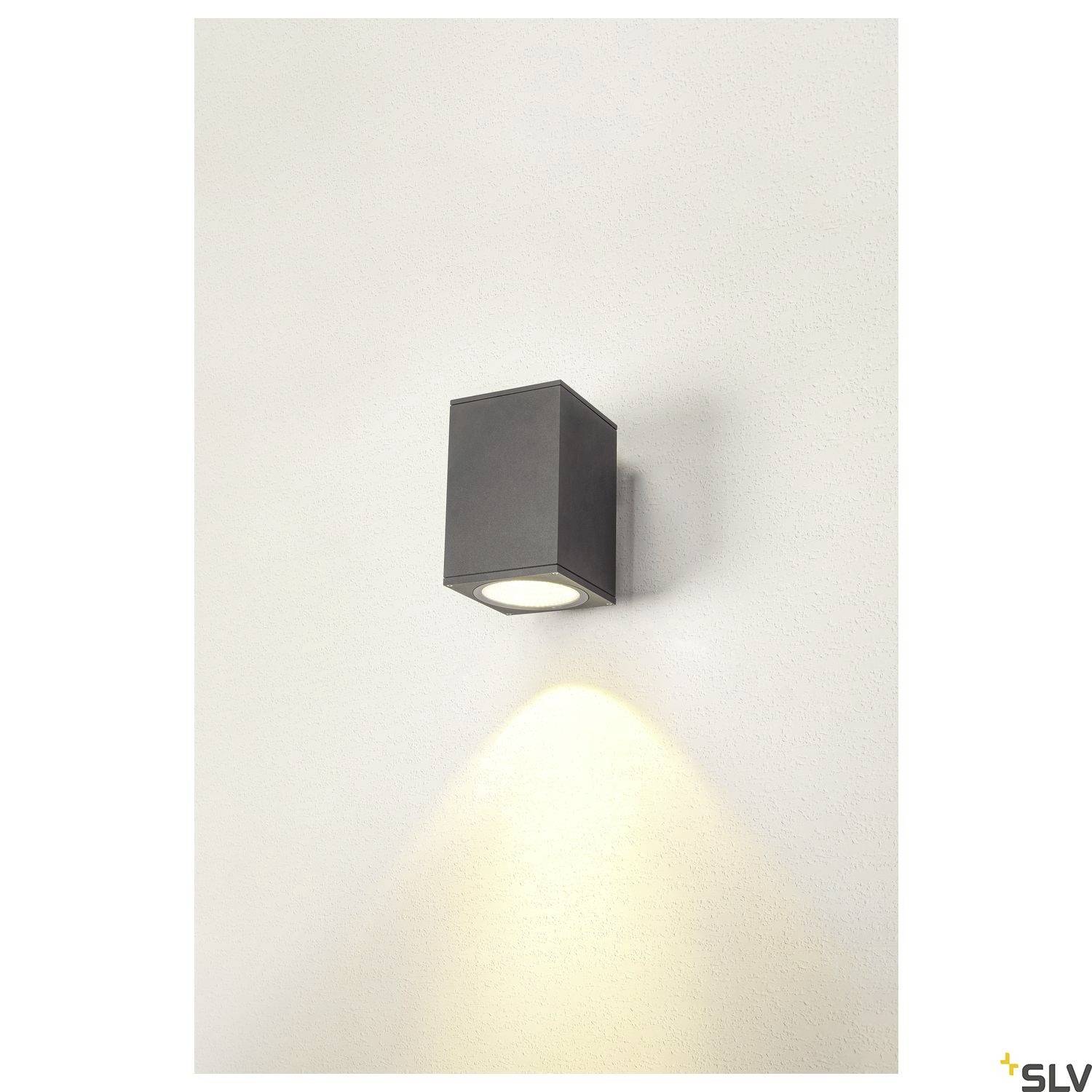 Wandlampe mit rechteckigem Gehäuse, aus dunklem Material. Leuchtet nach unten an einer weißen Wand.