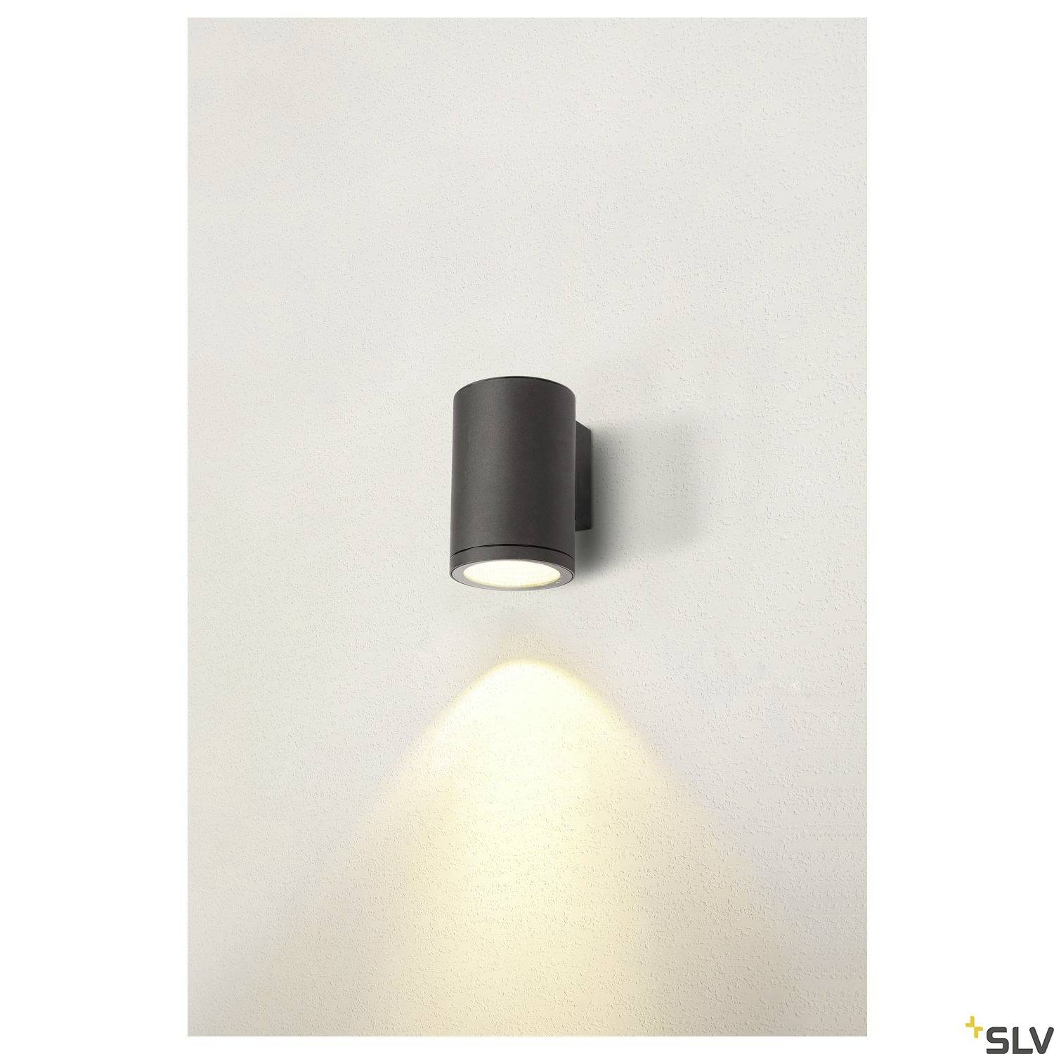 Wandleuchte in zylindrischer Form aus schwarzem Metall, wirft warmes Licht nach unten auf die weiße Wand.