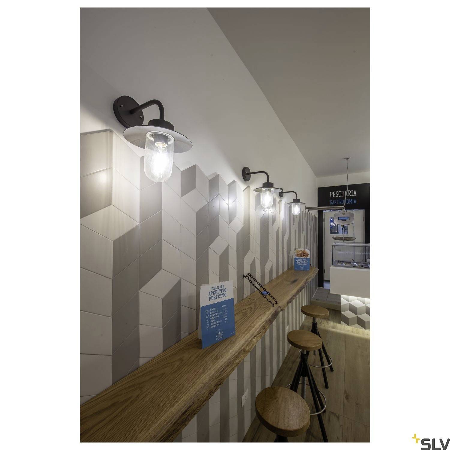 Ein moderner Tresen aus Holz mit Barhockern in einem Restaurant, an einer Wand mit geometrischen Fliesen und stilvollen Wandlampen.