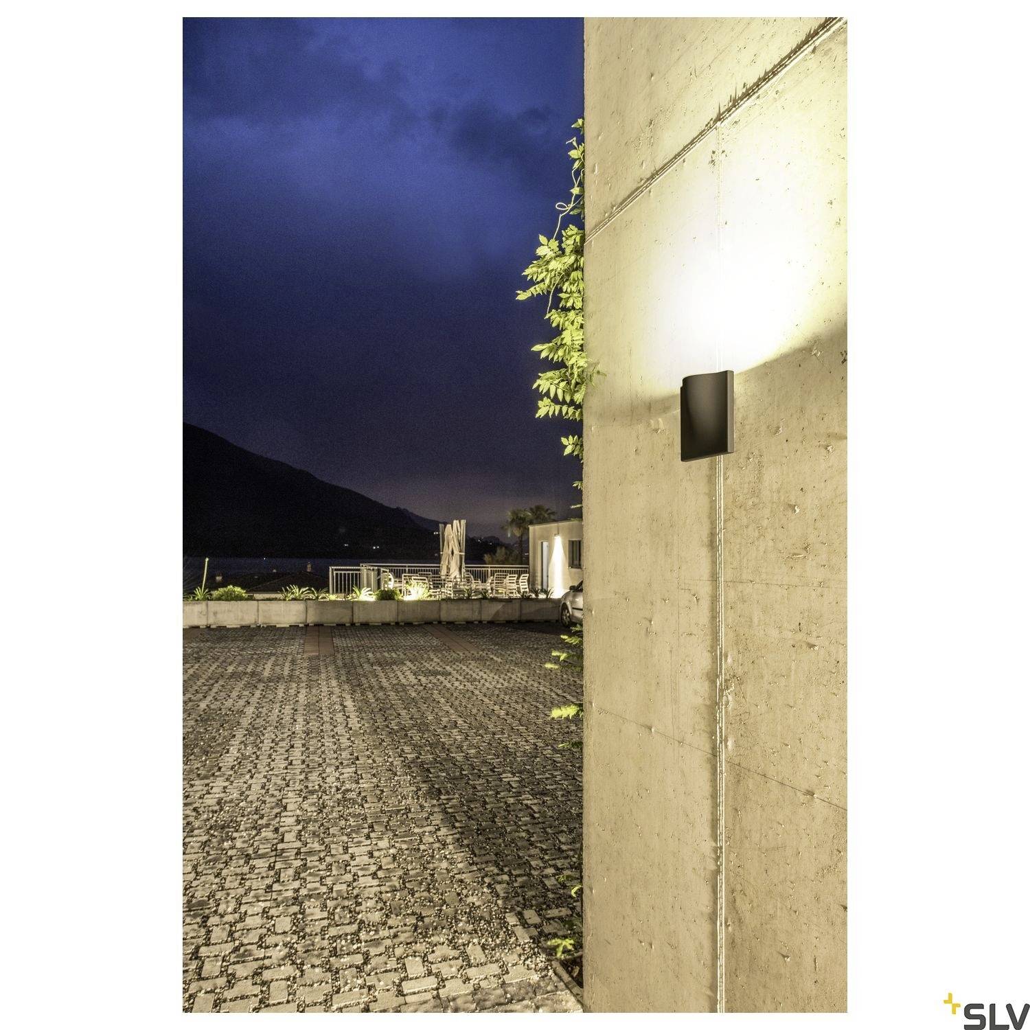 Außenmauer mit leuchtender Wandleuchte bei Dämmerung, beleuchtet gepflasterten Hof; Berge und Himmel im Hintergrund.
