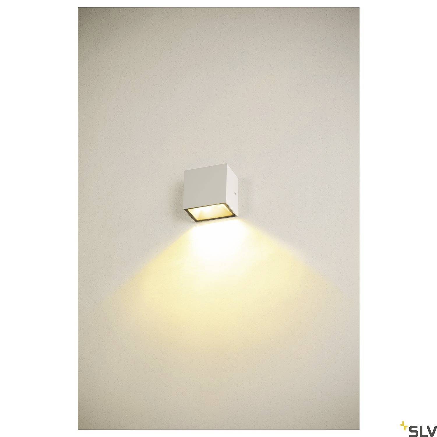 Weiße Wandleuchte wirft warmes Licht nach unten an eine beige Wand. Minimalistisches Design, geeignet für moderne Innenräume.