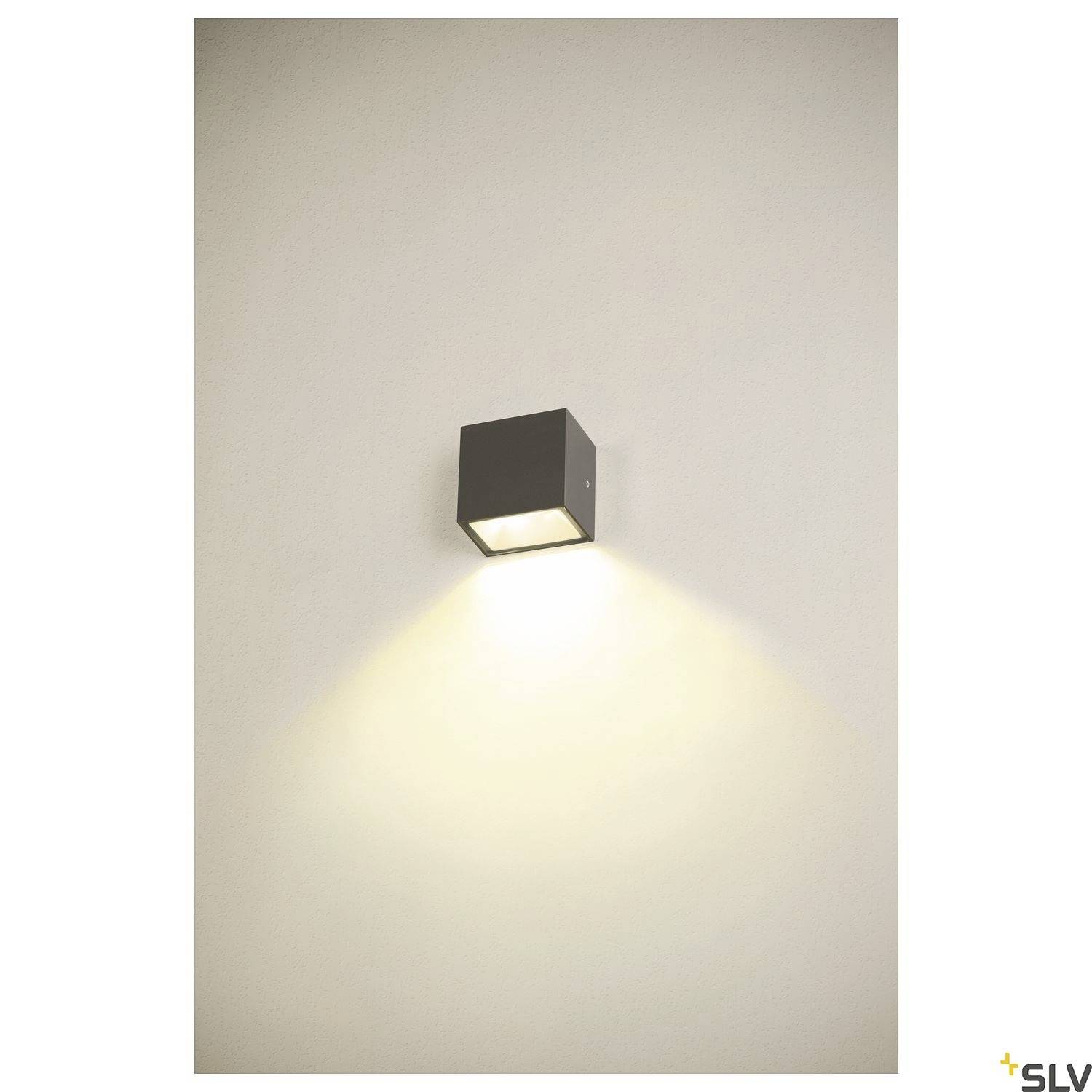 Schwarze Wandlampe in Würfelform strahlt warmes Licht nach unten auf eine Wand. Minimalistisches Design, geeignet für moderne Innenräume.