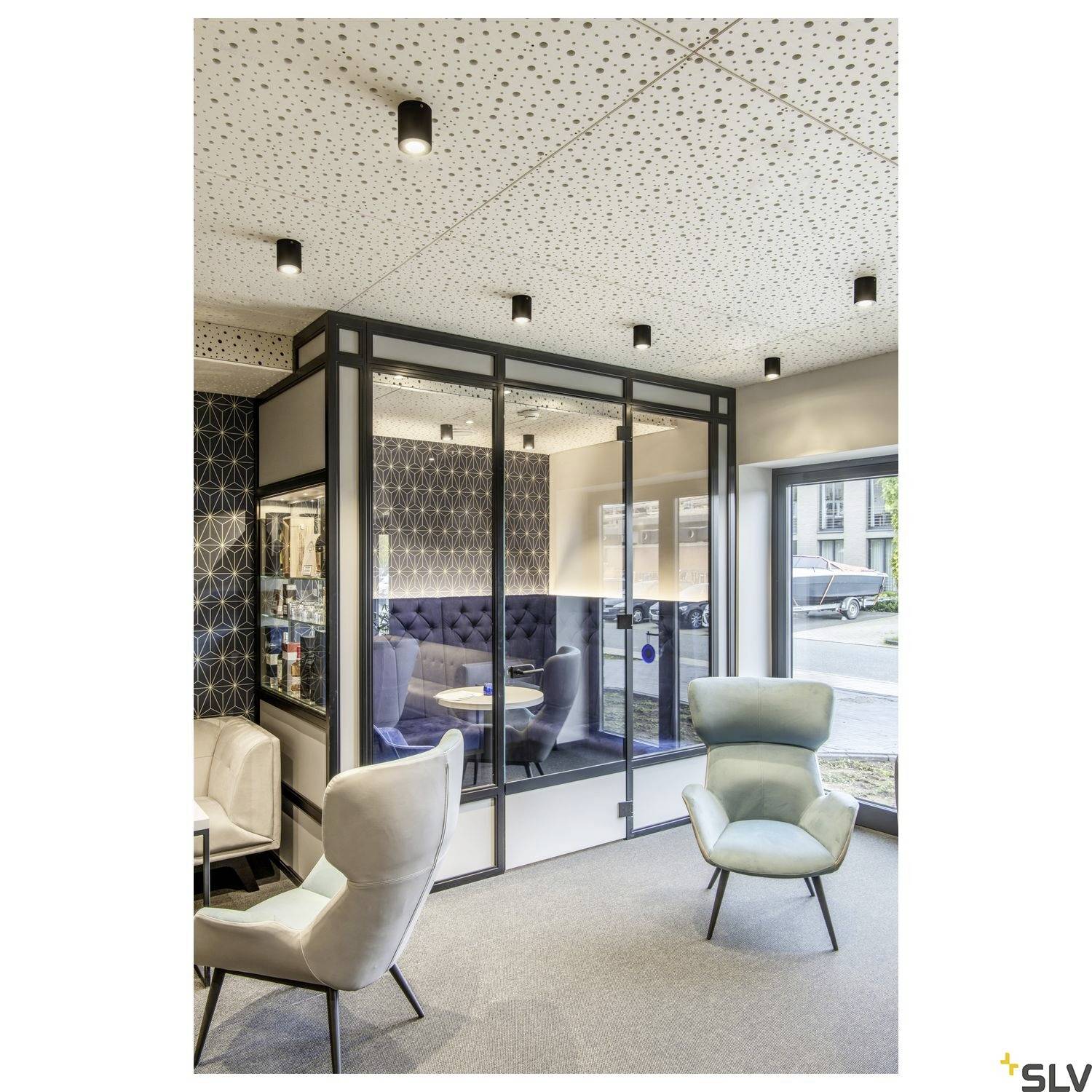 Ein modernes Büro mit Glasabtrennung, blaue Akzentstühle und Leuchten an der Decke. Fenster mit Blick nach draußen.