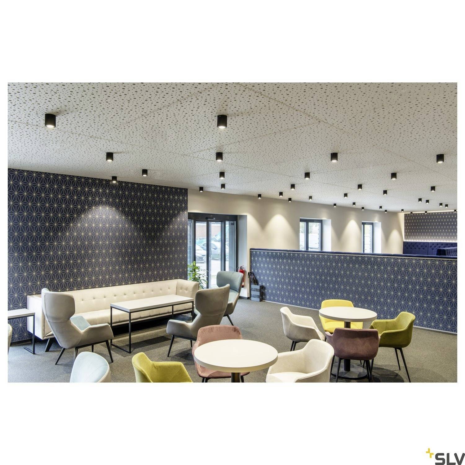 Moderne Lounge mit Sofas, Sessel in verschiedenen Farben und runden Tischen. Deckenleuchten und große Fenster im Hintergrund.