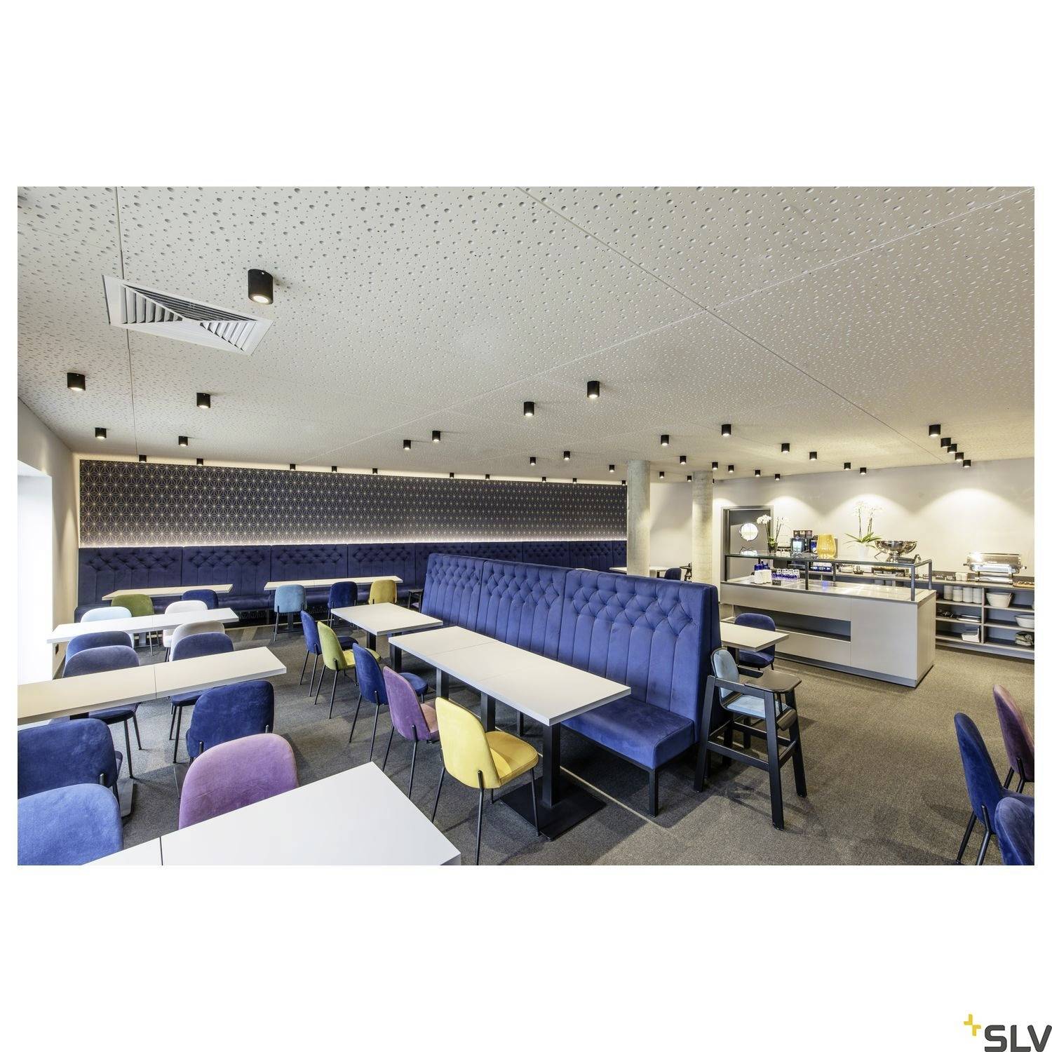 Ein modernes Café mit blauen und bunten Stühlen, weißen Tischen und einer Theke im Hintergrund. Deckenleuchten sorgen für Beleuchtung.