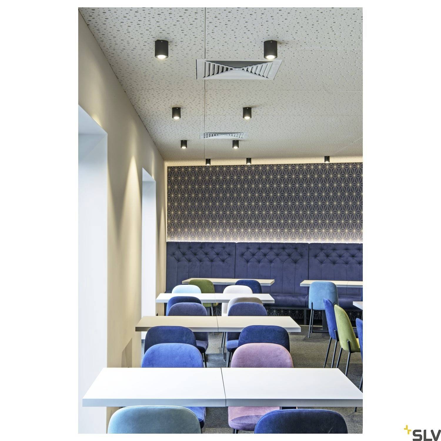 Kantine mit modernen blauen und rosa Stühlen und Tischen, beleuchtet durch Deckenspots; geometrisches Wandmuster im Hintergrund.