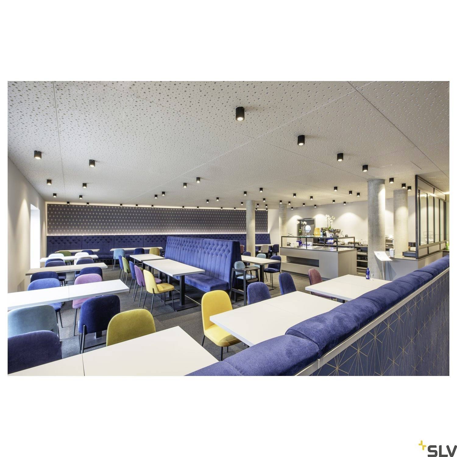 Großer, moderner Speiseraum mit weiß-gedeckten Tischen, bunten Stühlen und blauer Polsterbank. Deckenbeleuchtung sorgt für helles Ambiente.