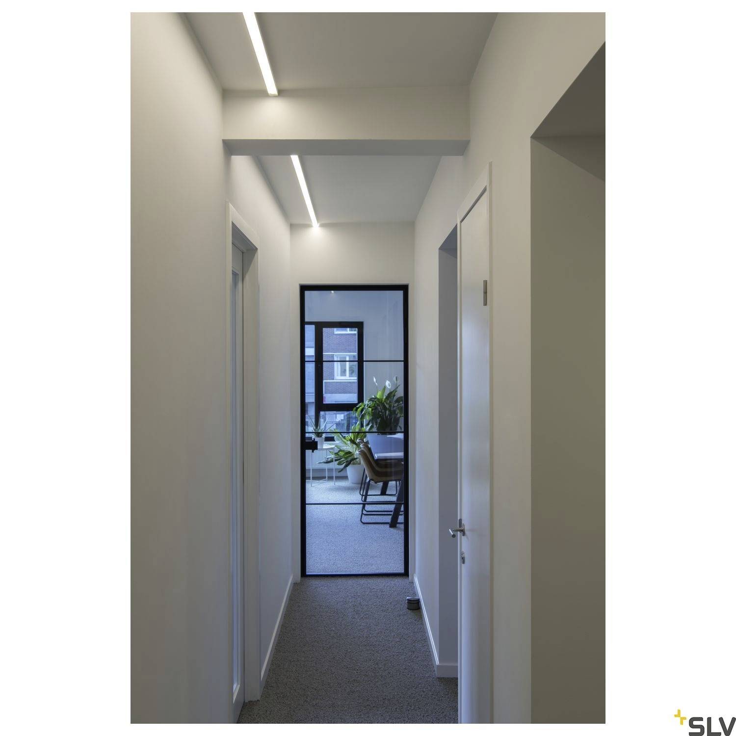 Ein langer, schmaler Flur mit weißen Wänden und einem Fenster am Ende, das Tageslicht in den Raum lässt. Eine Pflanze und Möbel sind im Fenster zu sehen.