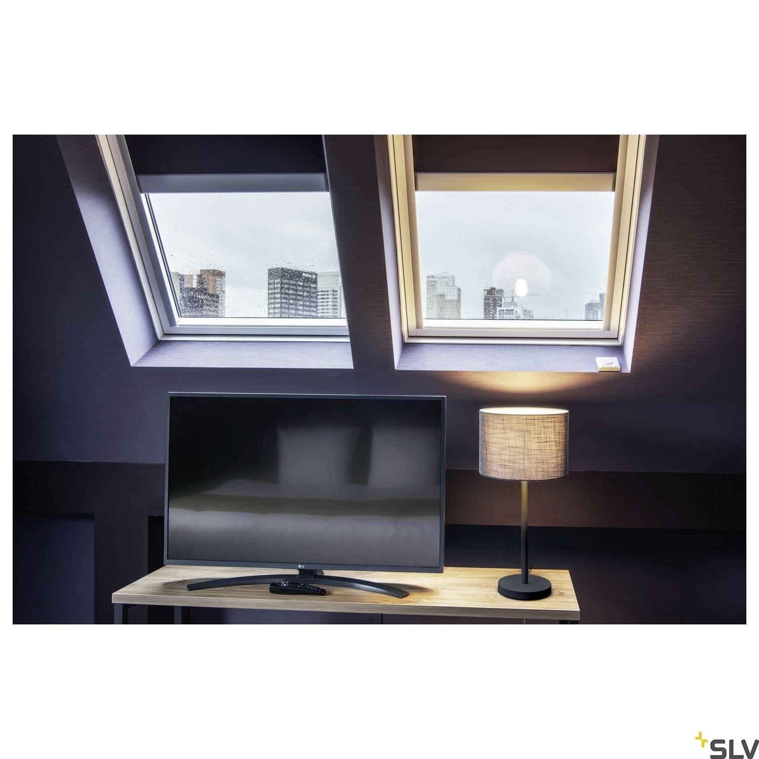 Ein Fernseher und eine Lampe stehen auf einem Tisch unter zwei Dachfenstern. Im Hintergrund sind verschwommene städtische Gebäude sichtbar.