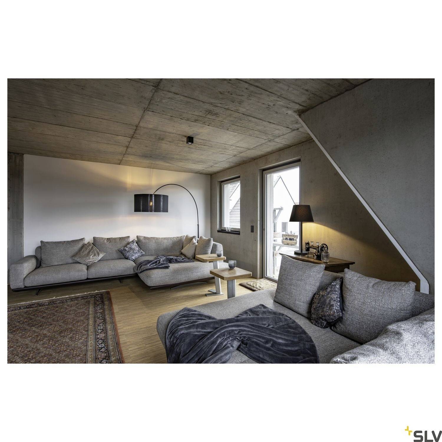 Gemütliches Wohnzimmer mit grauen Sofas, Holzboden, Stehlampe und einem Fenster rechts, das natürliches Licht hereinlässt.