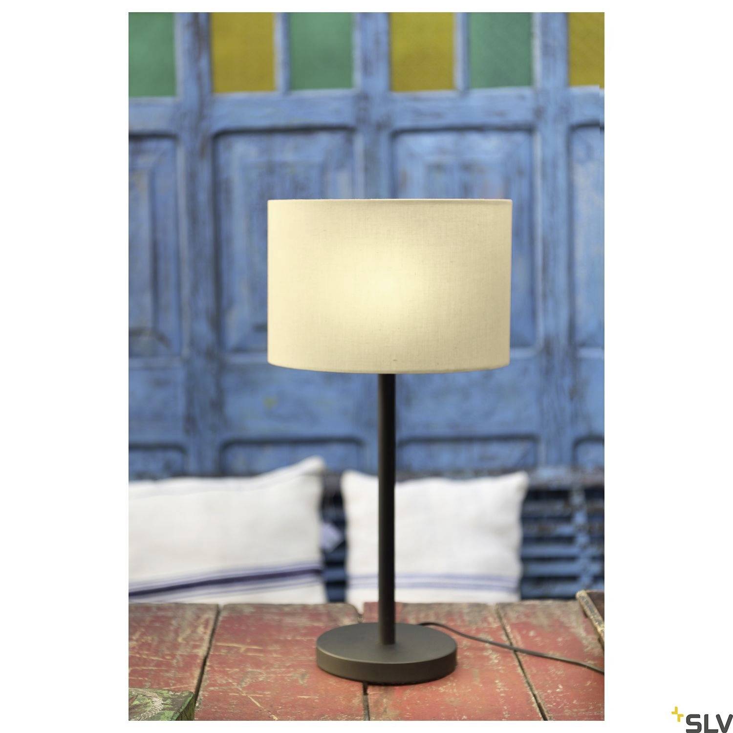 Eine Tischlampe mit hellem Lampenschirm steht auf einem Holztisch. Im Hintergrund sind blaue Holztüren und Kissen sichtbar.