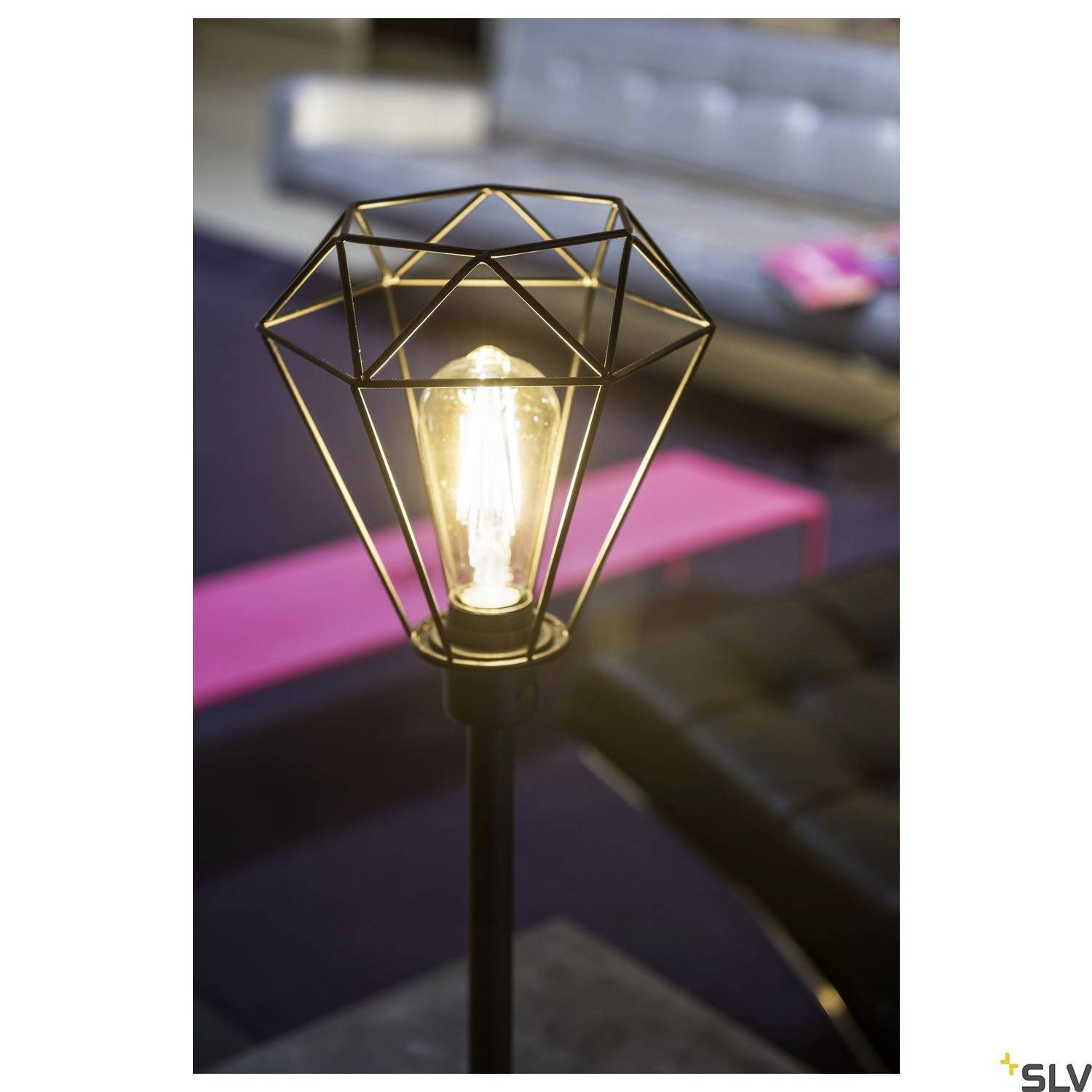 Eine moderne Stehlampe mit geometrischem, metallischem Lampenschirm leuchtet vor einem unscharfen Wohnzimmerhintergrund.