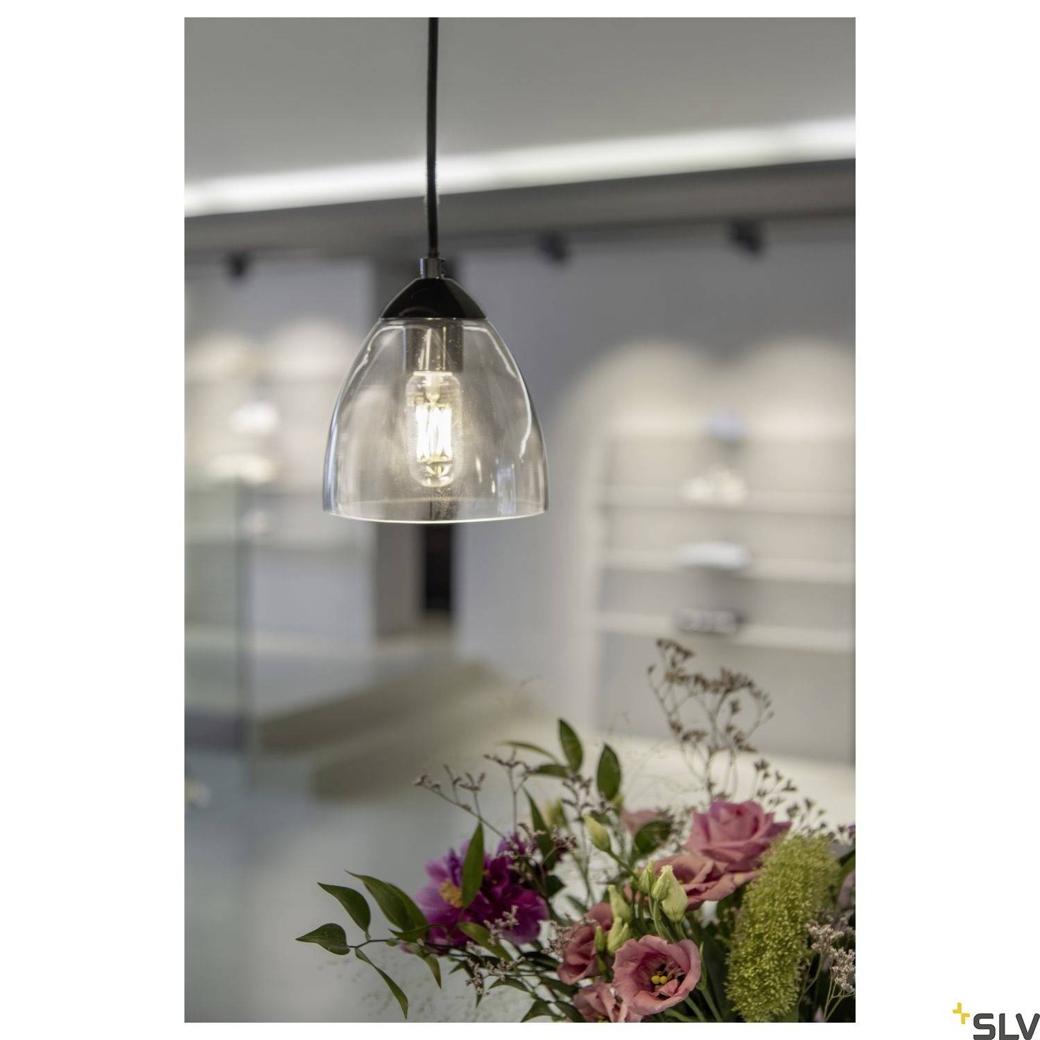Eine moderne Hängelampe beleuchtet einen Raum mit Regal im Hintergrund. Im Vordergrund ist ein Bouquet mit rosa und grünen Blumen zu sehen.