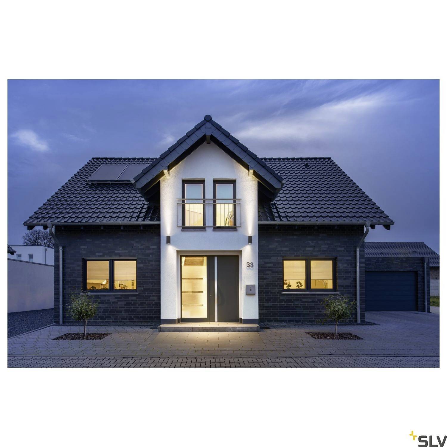 Einfamilienhaus bei Dämmerung mit eingeschalteten Außenleuchten, dunkelgraue Fassade, symmetrisch angeordnete Fenster und Dachgauben.