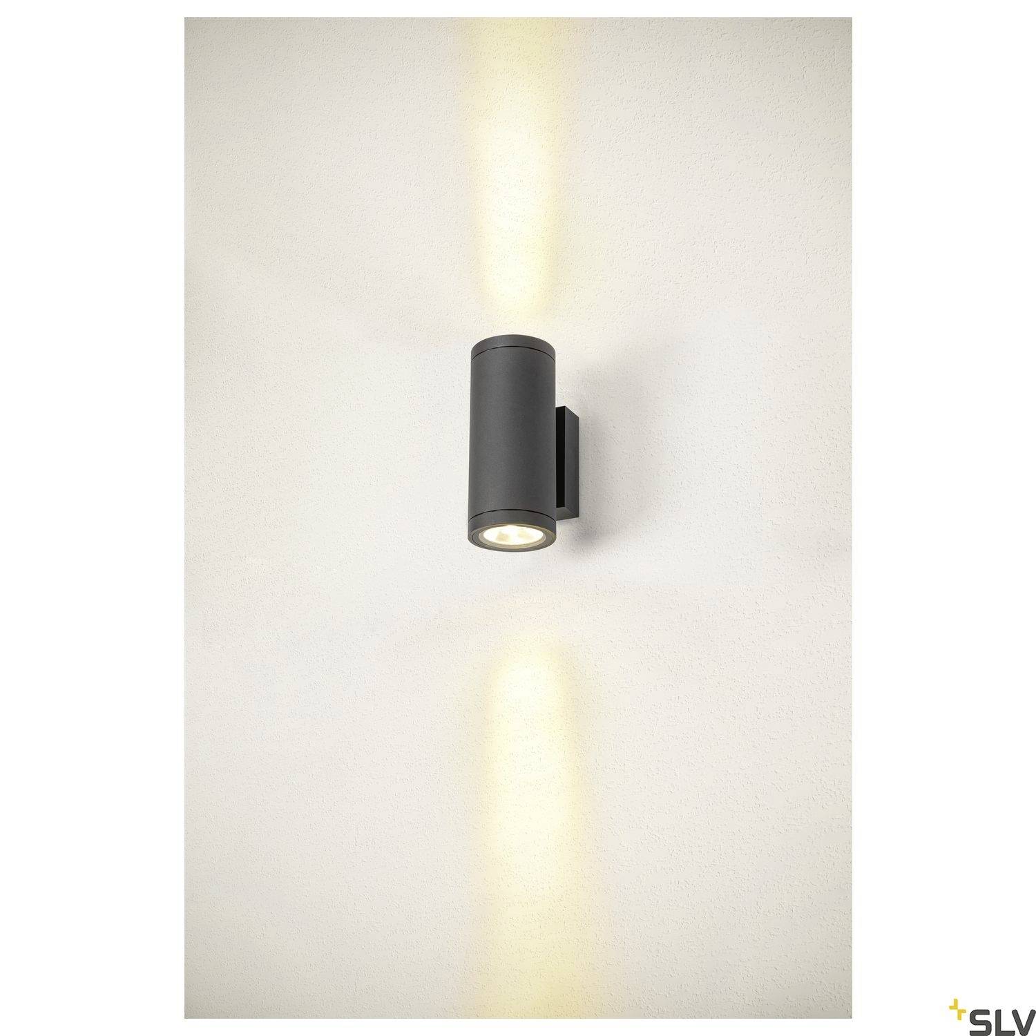 Eine zylindrische schwarze Wandleuchte strahlt sowohl nach oben als auch nach unten Licht aus, akzentuiert eine glatte, helle Wand.