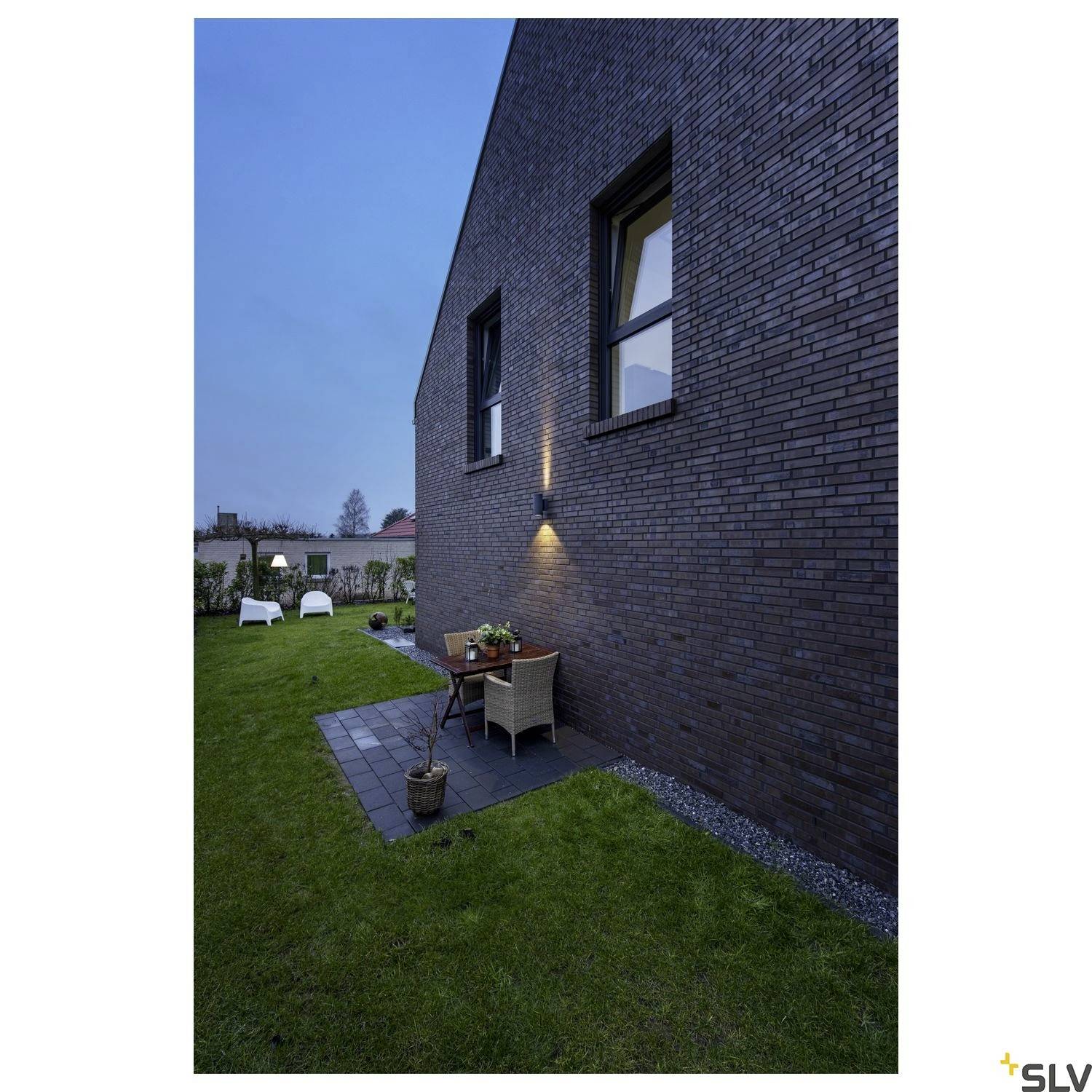 Moderne Außenansicht eines Ziegelhauses bei Dämmerung, beleuchtet von einer Außenlampe. Im Garten stehen weiße Stühle.
