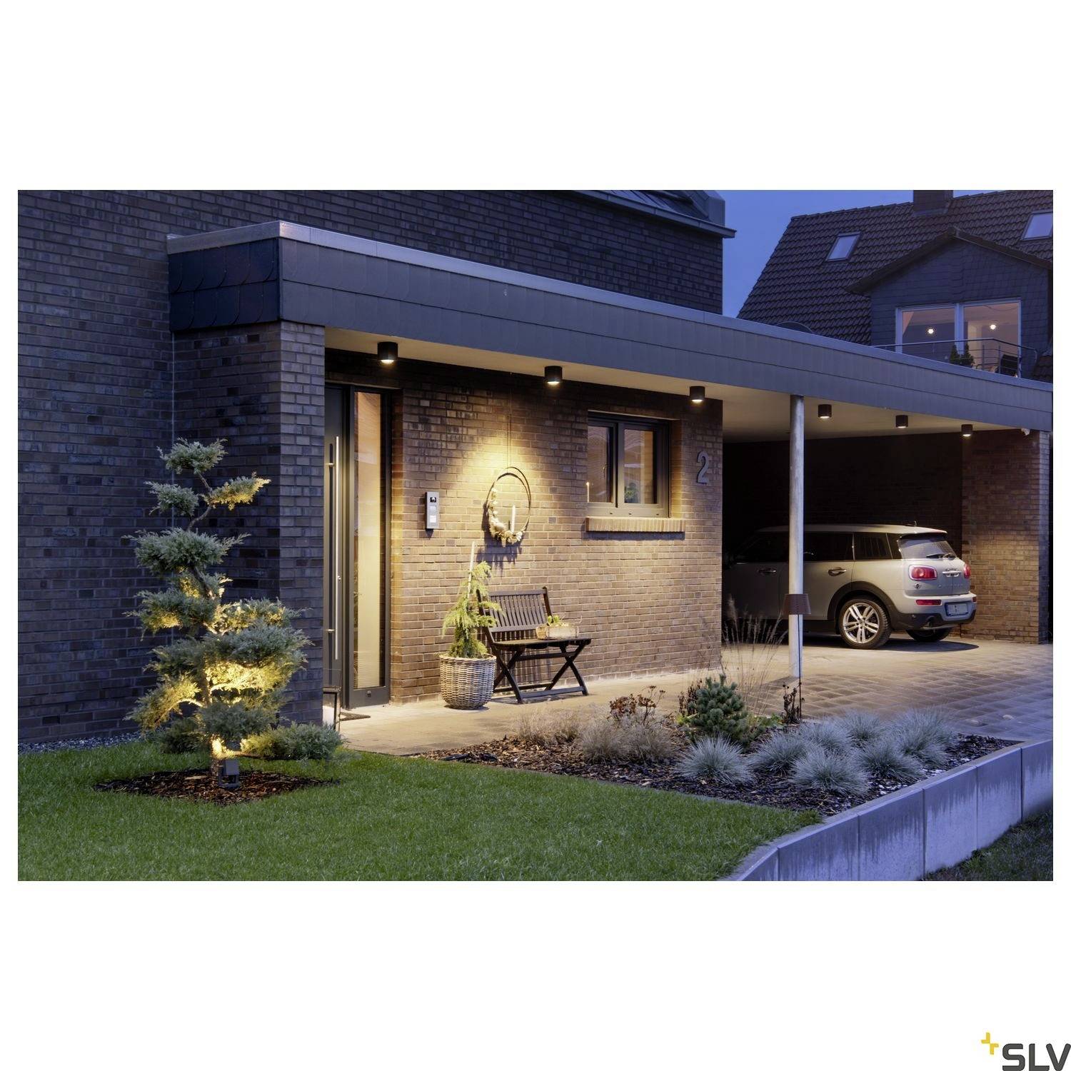 Modernes Haus mit Backsteinfassade, beleuchtetem Eingang und Carport. Vor dem Eingang stehen ein beleuchteter Baum und Pflanzen.
