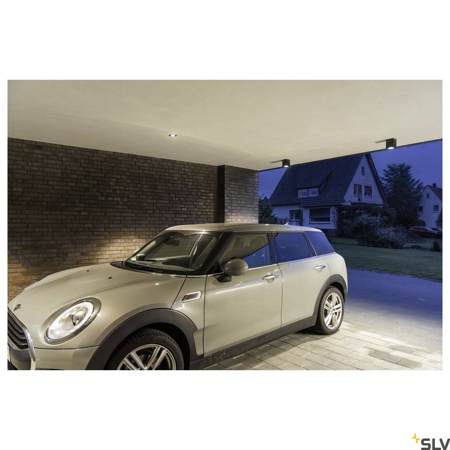 Auto steht in moderner Garage neben gemauerter Wand. Außen ist ein Wohngebiet mit sichtbaren Häusern und Rasen im Hintergrund.