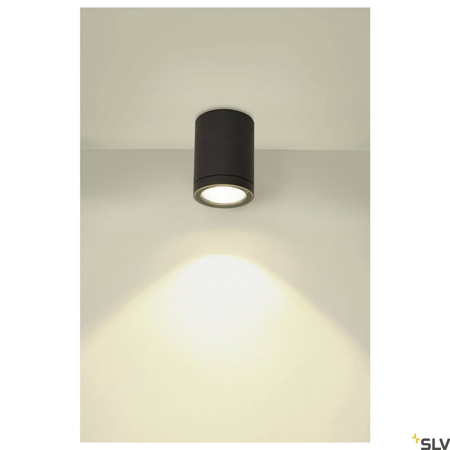 Eine zylindrische schwarze Deckenleuchte strahlt gleichmäßiges Licht auf die Wand aus, wodurch ein sanfter Lichtkegel entsteht.