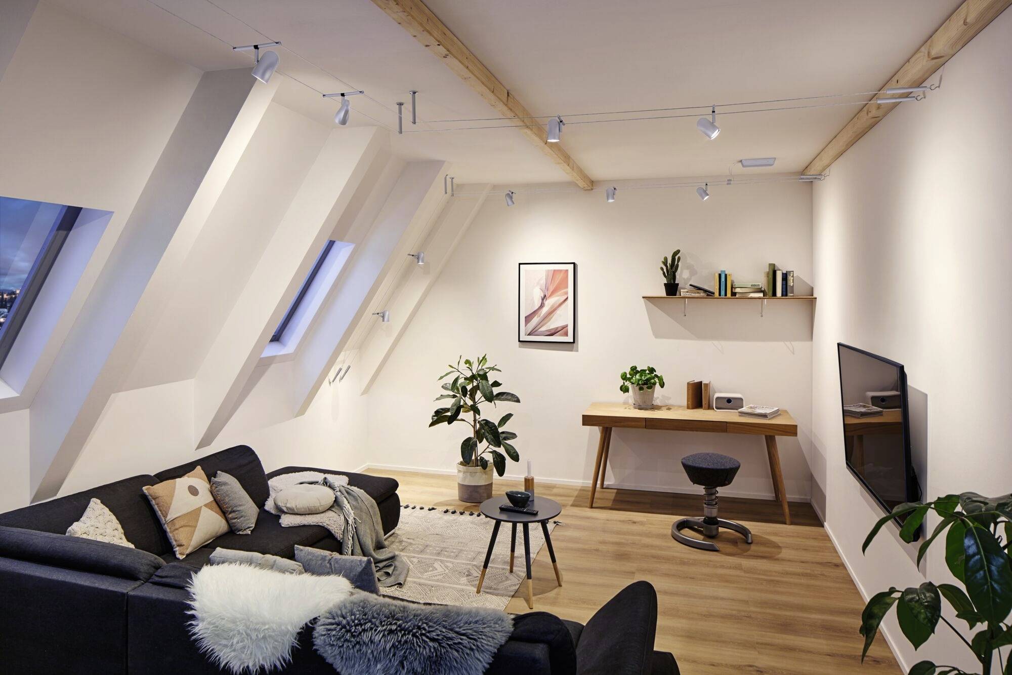 Gemütliches Wohnzimmer in einer Dachgeschosswohnung mit Sofa, Pflanzen, Schreibtisch und Fernseher. Moderne Beleuchtung an der Decke.
