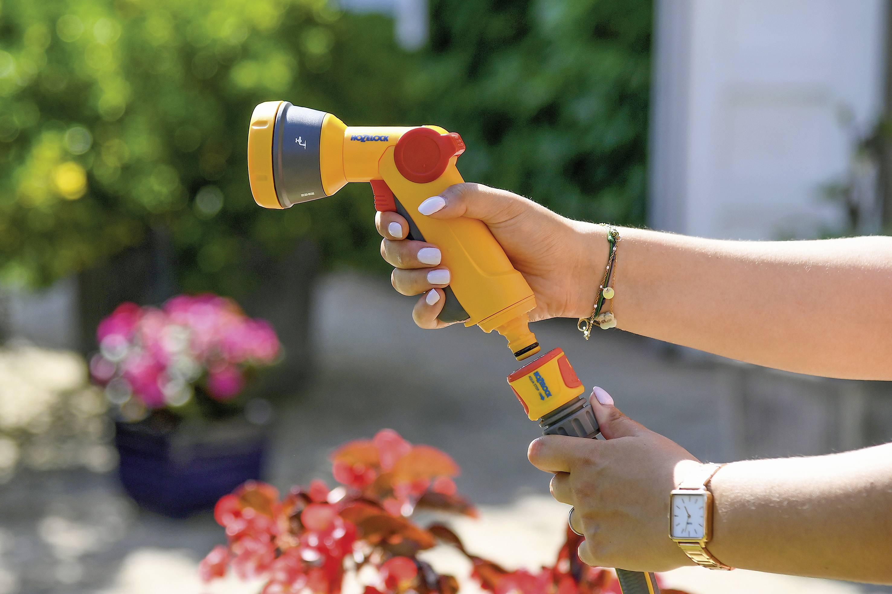Jemand hält einen gelben Gartenschlauch mit Sprühaufsatz in der Hand, bereit zum Gießen, mit blühenden Pflanzen im Hintergrund.