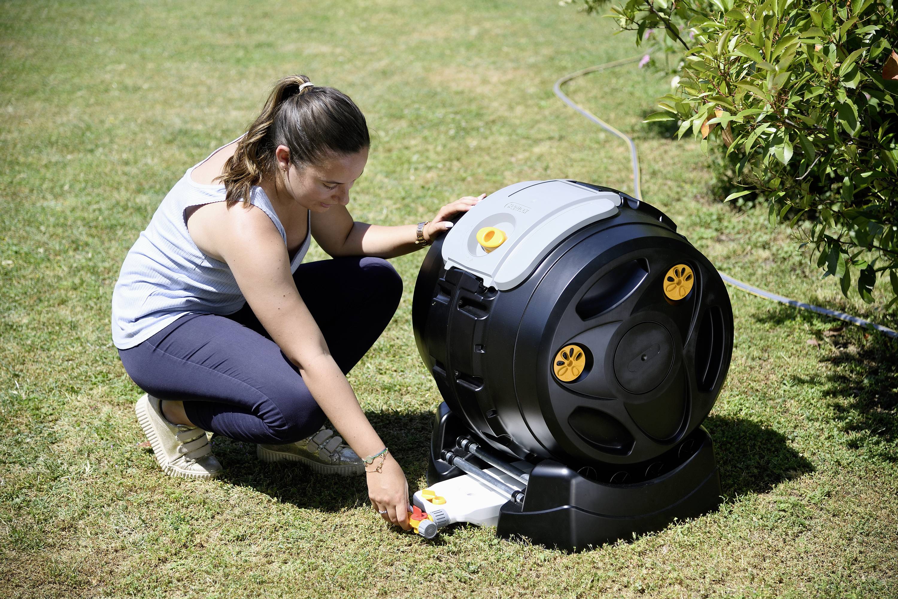 Eine Frau kniet auf dem Rasen und bedient einen schwarzen, runden Gartenschlauch-Aufroller. Im Hintergrund sind Pflanzen zu sehen.