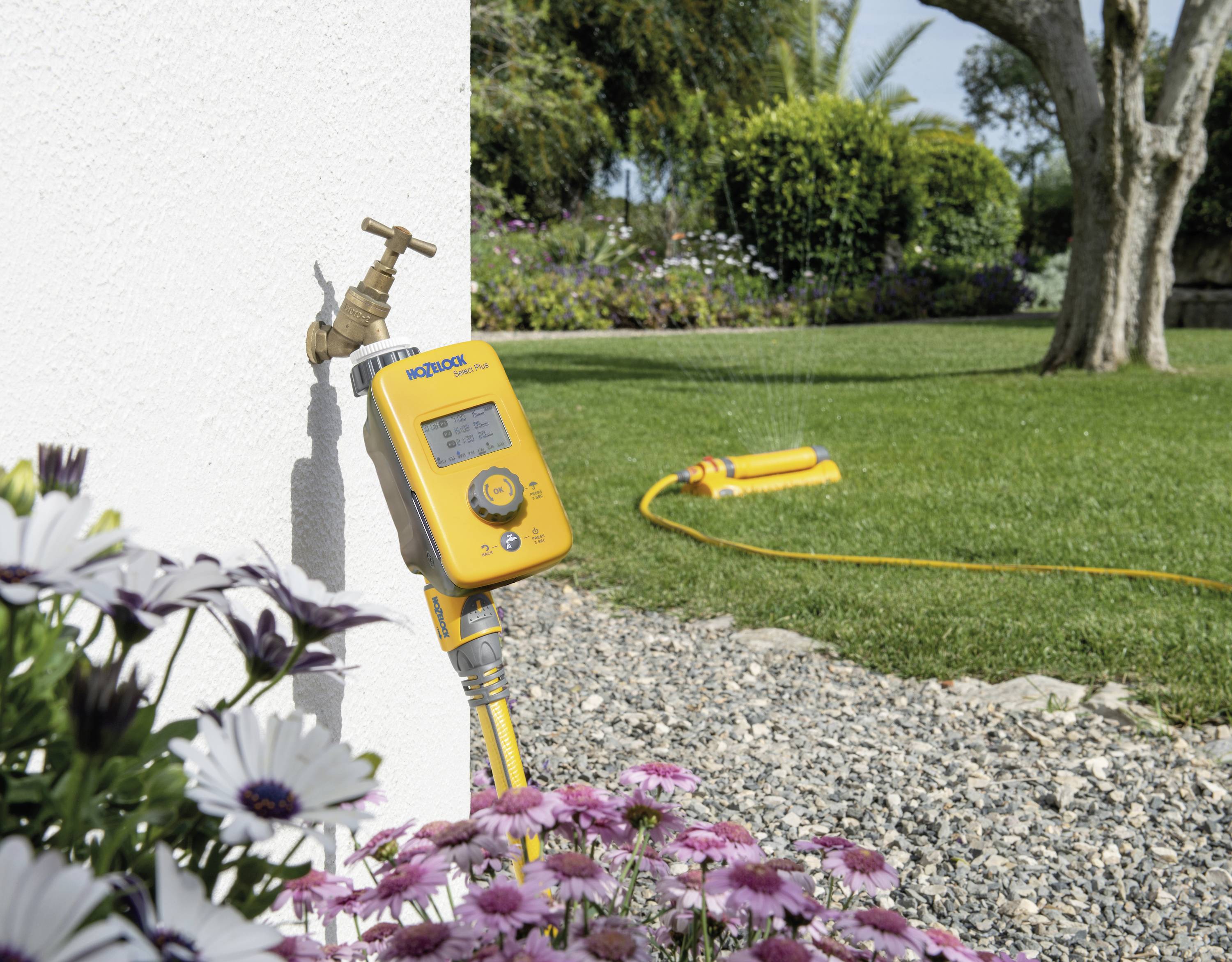 Ein gelber Gartenbewässerungstimer ist an einen Wasserhahn angeschlossen, während im Hintergrund ein Rasensprenger in einem Garten läuft.