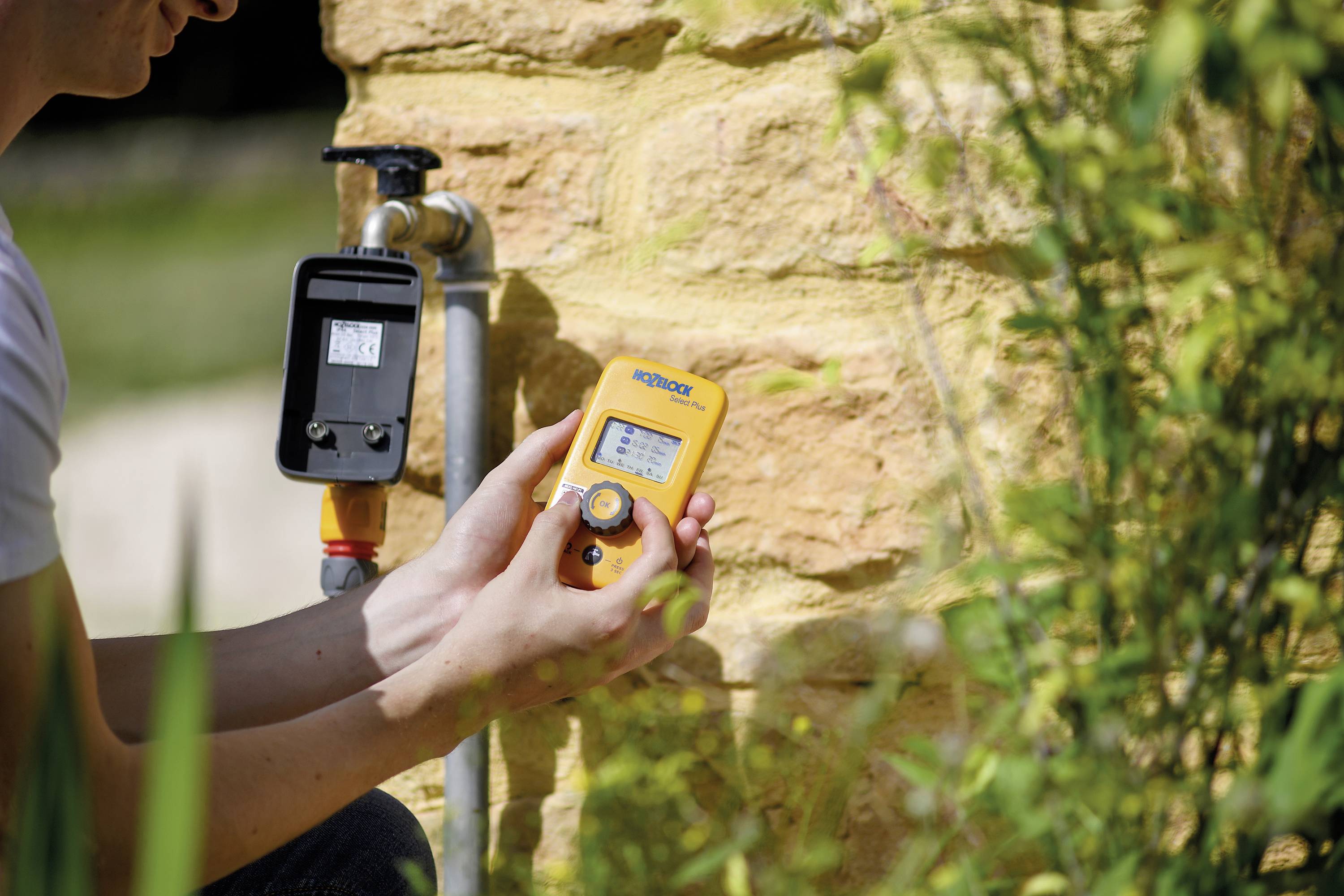 Eine Person hält ein gelbes digitales Messgerät nahe eines Wasserhahns an einer Steinmauer. Pflanzenrahmen das Bild ein.