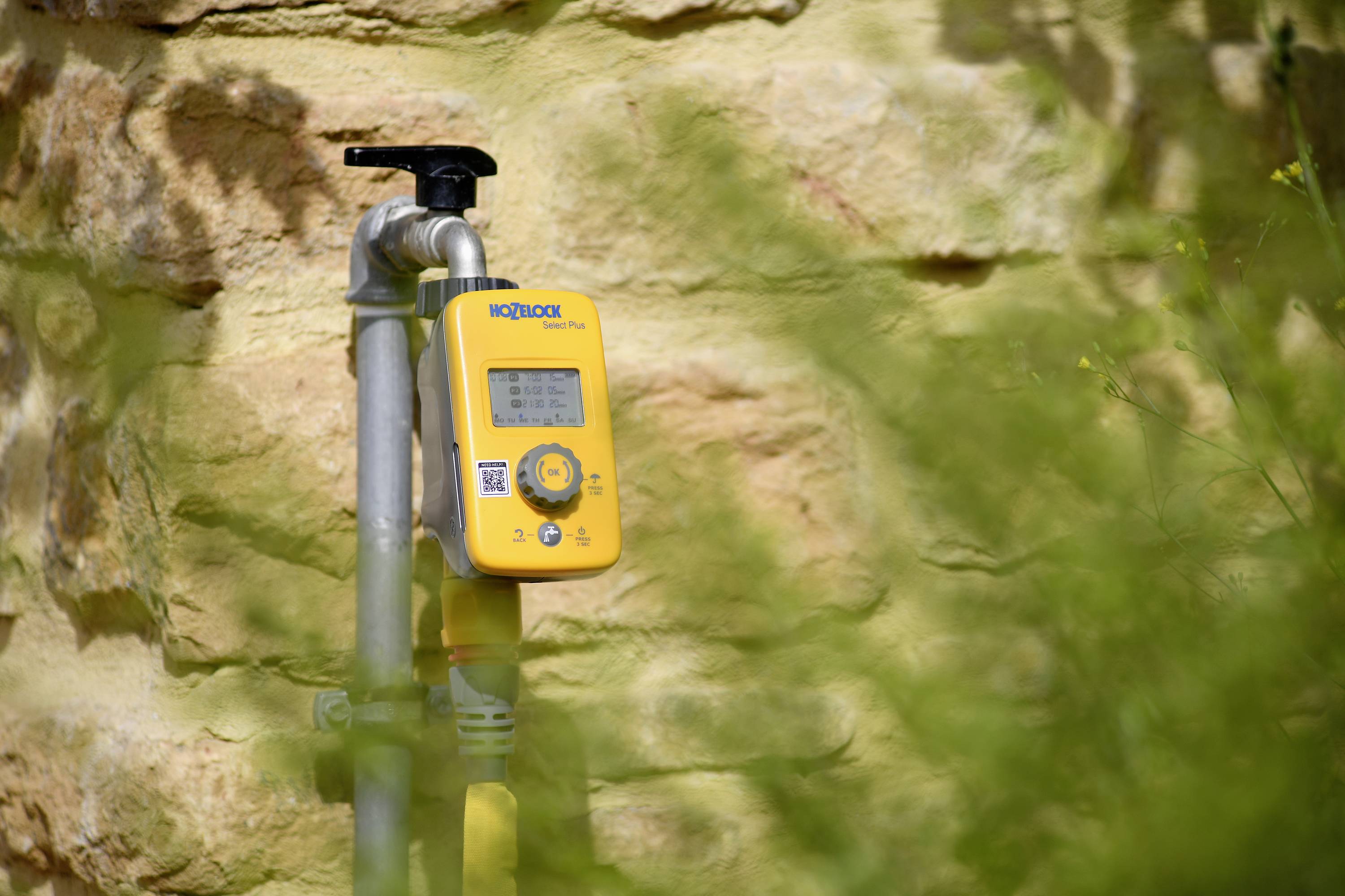Ein gelbes Bewässerungssteuergerät ist an einem Rohr vor einer beige-gelben Steinwand befestigt, umgeben von unscharfer Vegetation.
