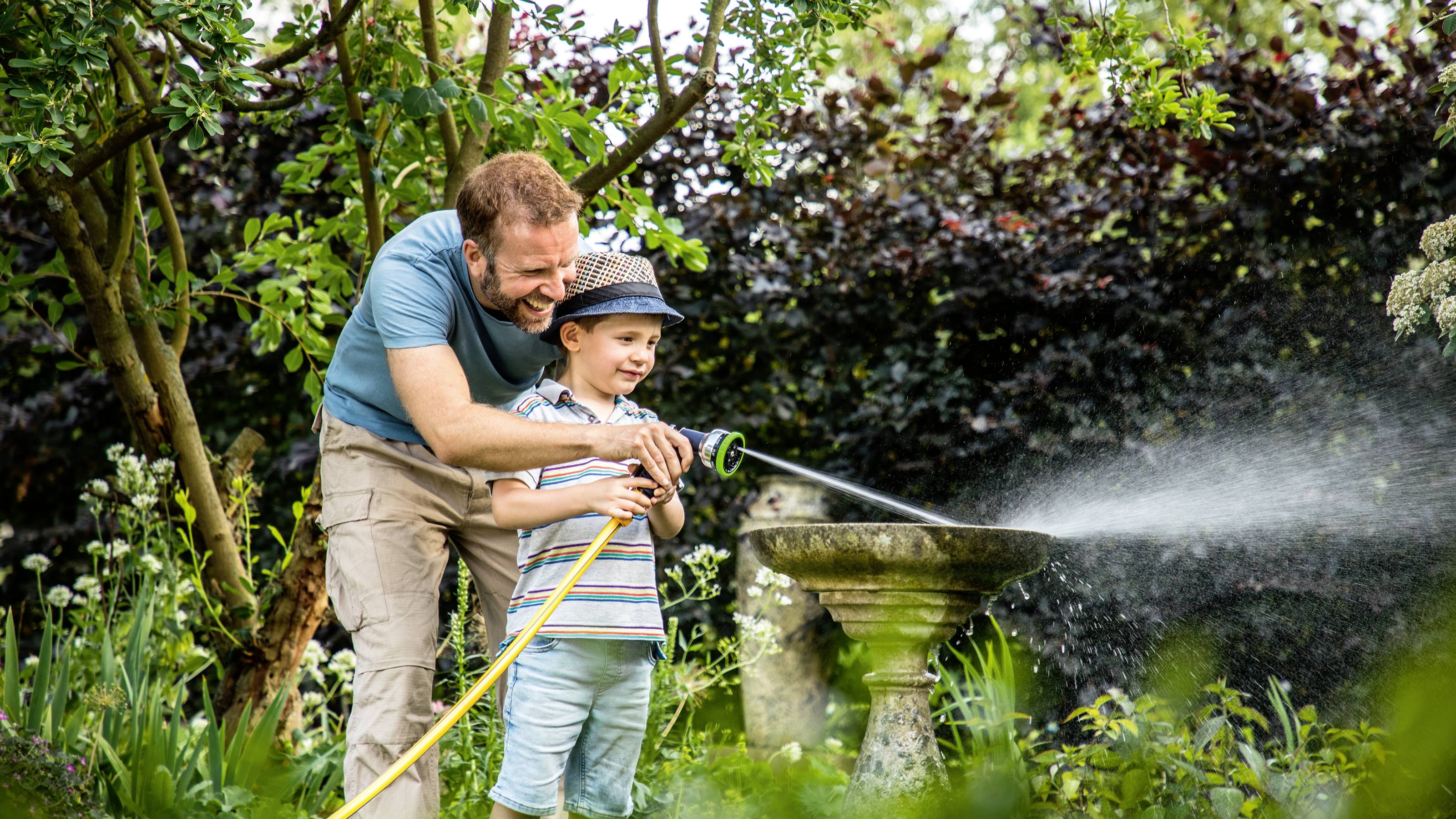 Ein Mann hilft einem Kind, mit einem Gartenschlauch in einem blühenden Garten Wasser zu sprühen, während sie beide lächeln.