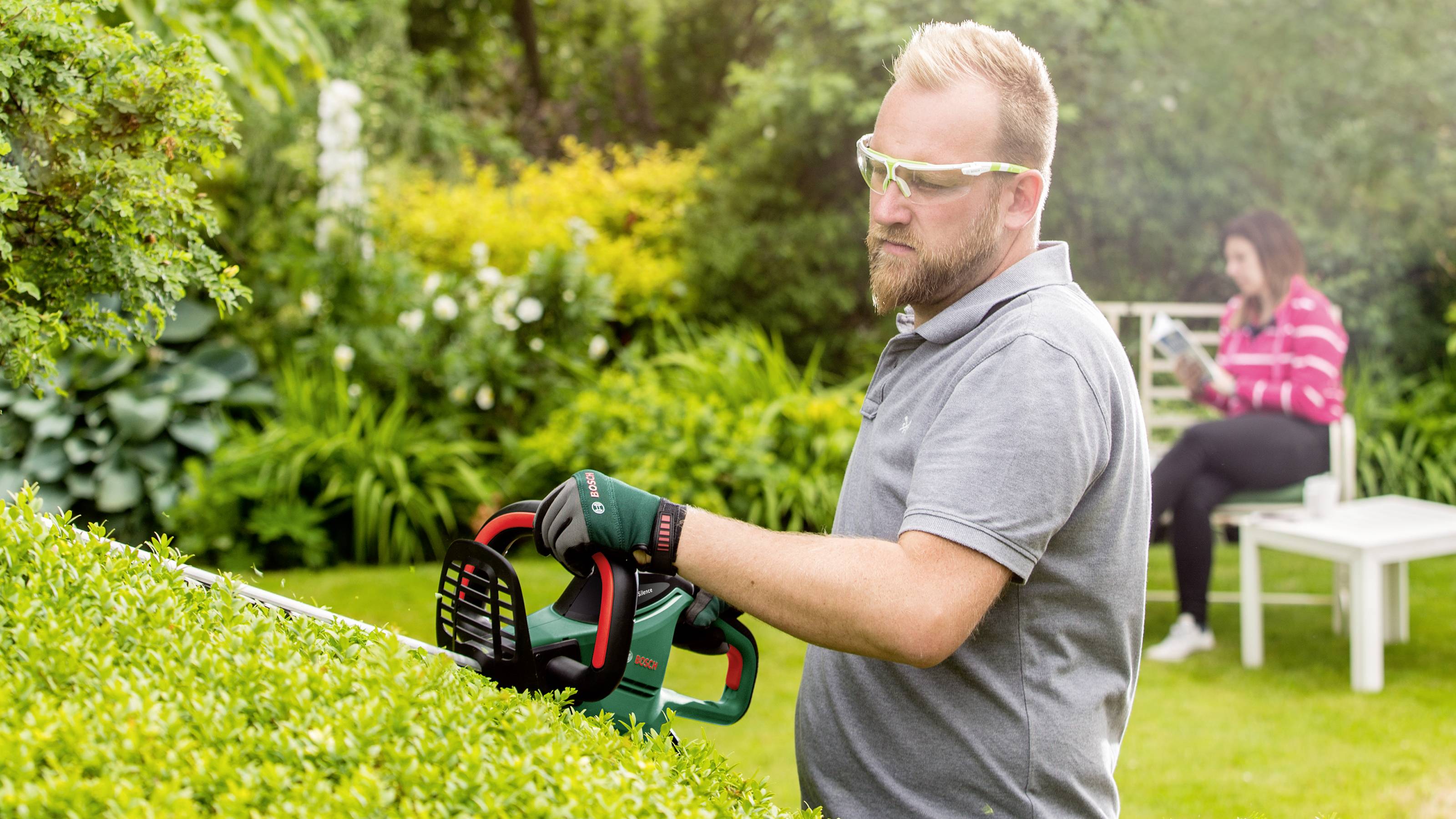 Ein Mann mit Schutzbrille und Handschuhen schneidet mit einer elektrischen Heckenschere eine Hecke im Garten. Im Hintergrund sitzt eine weitere Person auf einer Bank.