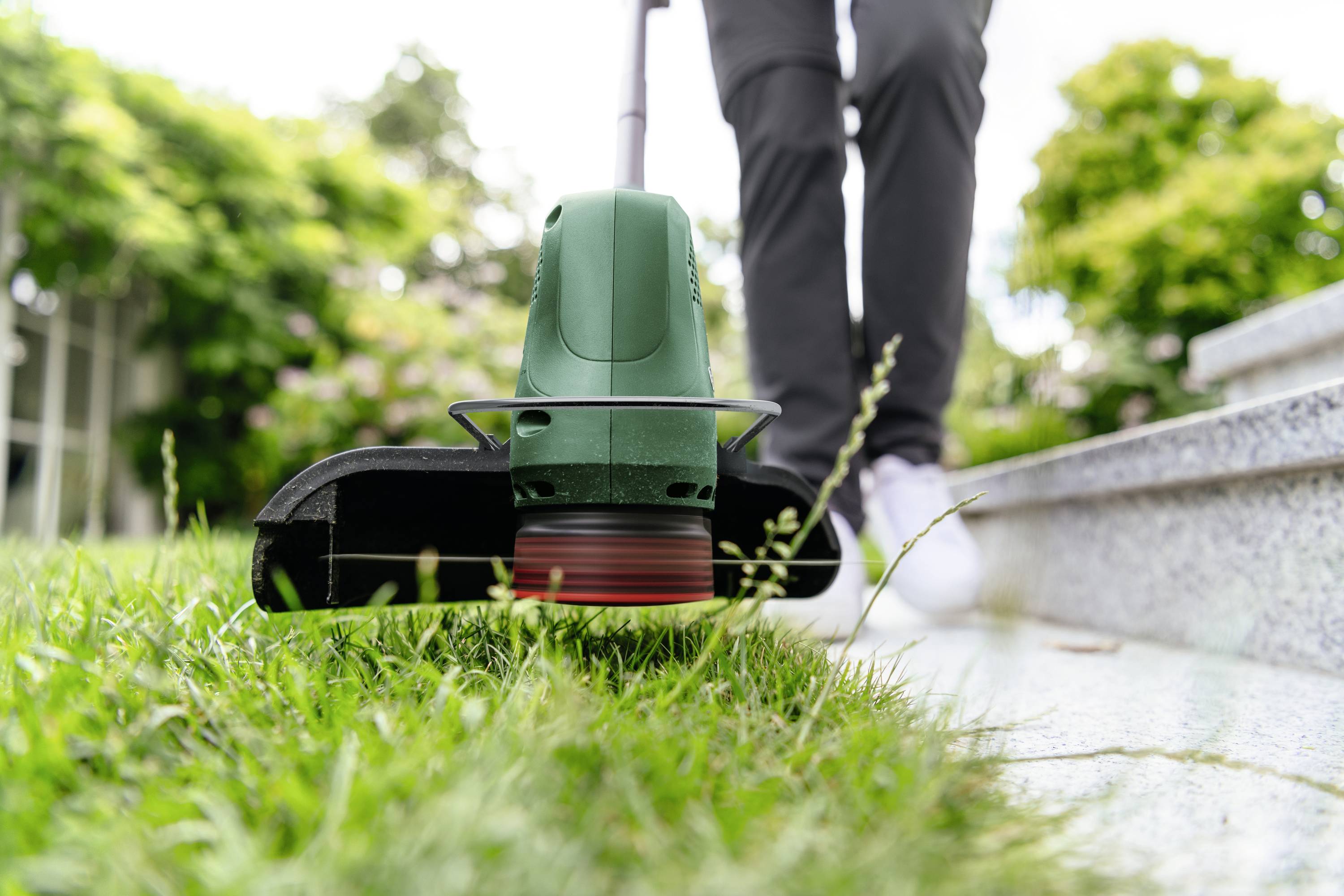 Person benutzt einen grünen Rasentrimmer, um Gras an einer Pflasterkante zu schneiden. Umgebung ist ein gepflegter Garten.