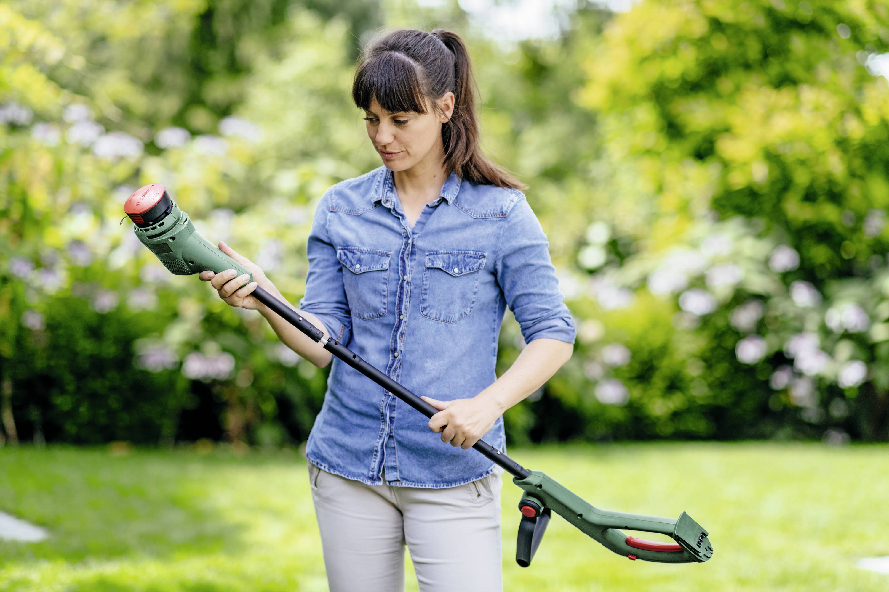 Eine Frau in einem Park hält einen elektrischen Rasentrimmer und inspiziert ihn sorgfältig. Im Hintergrund grüne Bäume und Sträucher.