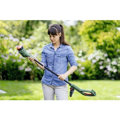 Eine Frau in einem Park hält einen elektrischen Rasentrimmer und inspiziert ihn sorgfältig. Im Hintergrund grüne Bäume und Sträucher.