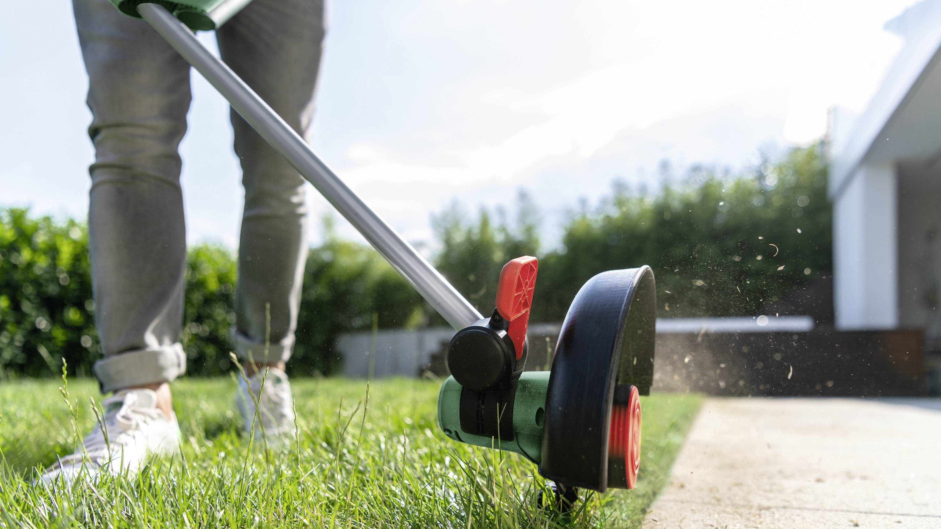 Person nutzt Rasentrimmer, um Gras an einer Gehwegkante zu trimmen. Graspartikel spritzen, und im Hintergrund sieht man einen Garten.