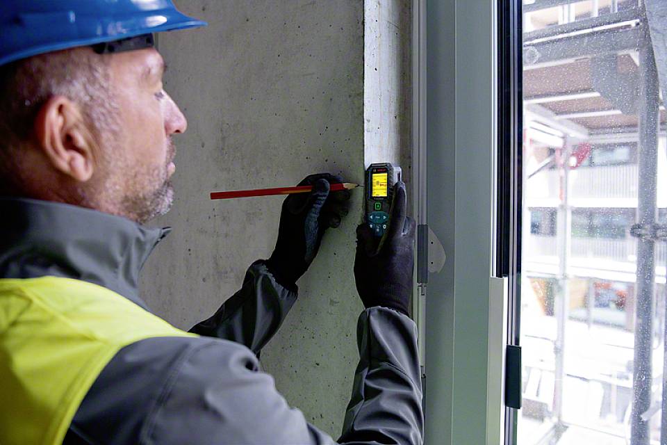 Ein Bauarbeiter misst eine Fensteröffnung mit einem digitalen Messgerät, um genaue Werte zu erhalten. Er trägt Schutzkleidung und Helm.