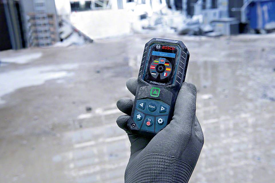 Eine Hand hält ein Laser-Entfernungsmesser auf einer Baustelle. Das Gerät zeigt Messwerte an, während im Hintergrund unscharfe Baustellenmaterialien zu sehen sind.