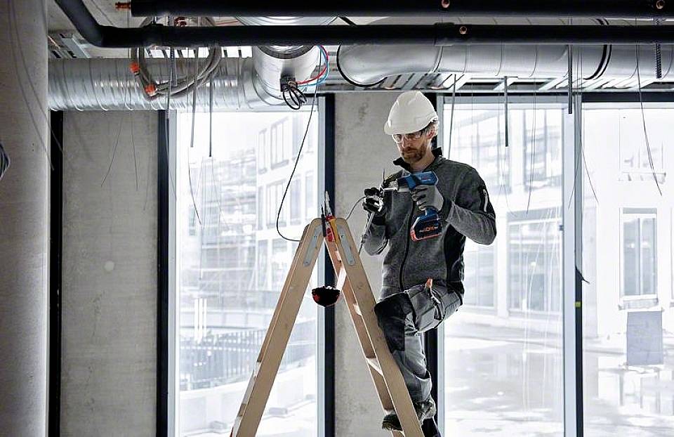 Ein Bauarbeiter mit Helm und Schutzbrille steht auf einer Leiter in einem modernen Gebäude und bohrt in die Decke.