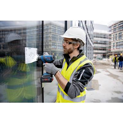 Ein Bauarbeiter mit Schutzhelm und Warnweste schraubt ein Papier mit einer Bohrmaschine an eine Glaswand auf einer Baustelle fest.