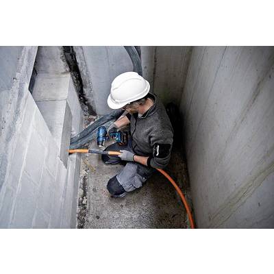 Ein Arbeiter mit Helm bohrt ein Loch in eine Betonwand, während er ein orangefarbenes Kabel durchzieht. Er sitzt in einem engen Raum.