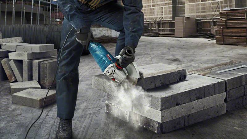 Ein Arbeiter schneidet mit einer elektrischen Säge Betonplatten auf einer Baustelle. Staub steigt auf, der Boden ist mit Platten bedeckt.