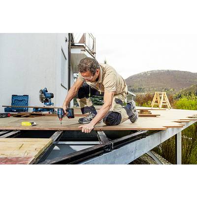 Ein Handwerker baut eine Holzterrasse an einem Haus. Er trägt Schutzbrille und benutzt einen Bohrer. Im Hintergrund sind Bäume und ein Hügel zu sehen.