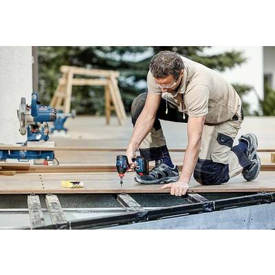 Ein Handwerker kniet auf einer Terrasse und befestigt Holzplanken mit einem Akkuschrauber. Im Hintergrund sind Werkzeuge sichtbar.