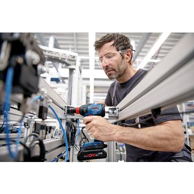Ein Mann in Schutzbrille arbeitet mit einem Akkuschrauber an einem industriellen Montageprojekt, umgeben von Metallgestellen und Kabeln.