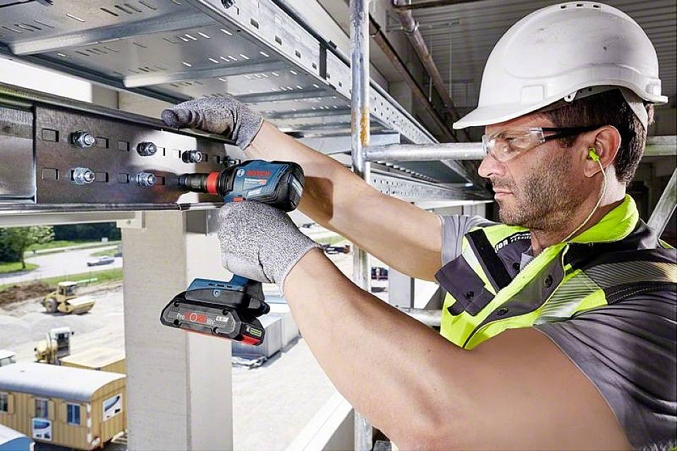 Ein Bauarbeiter in Schutzkleidung nutzt einen Akku-Bohrschrauber, um Metallstreben an einem Gebäude zu befestigen. Im Hintergrund Baustelle.