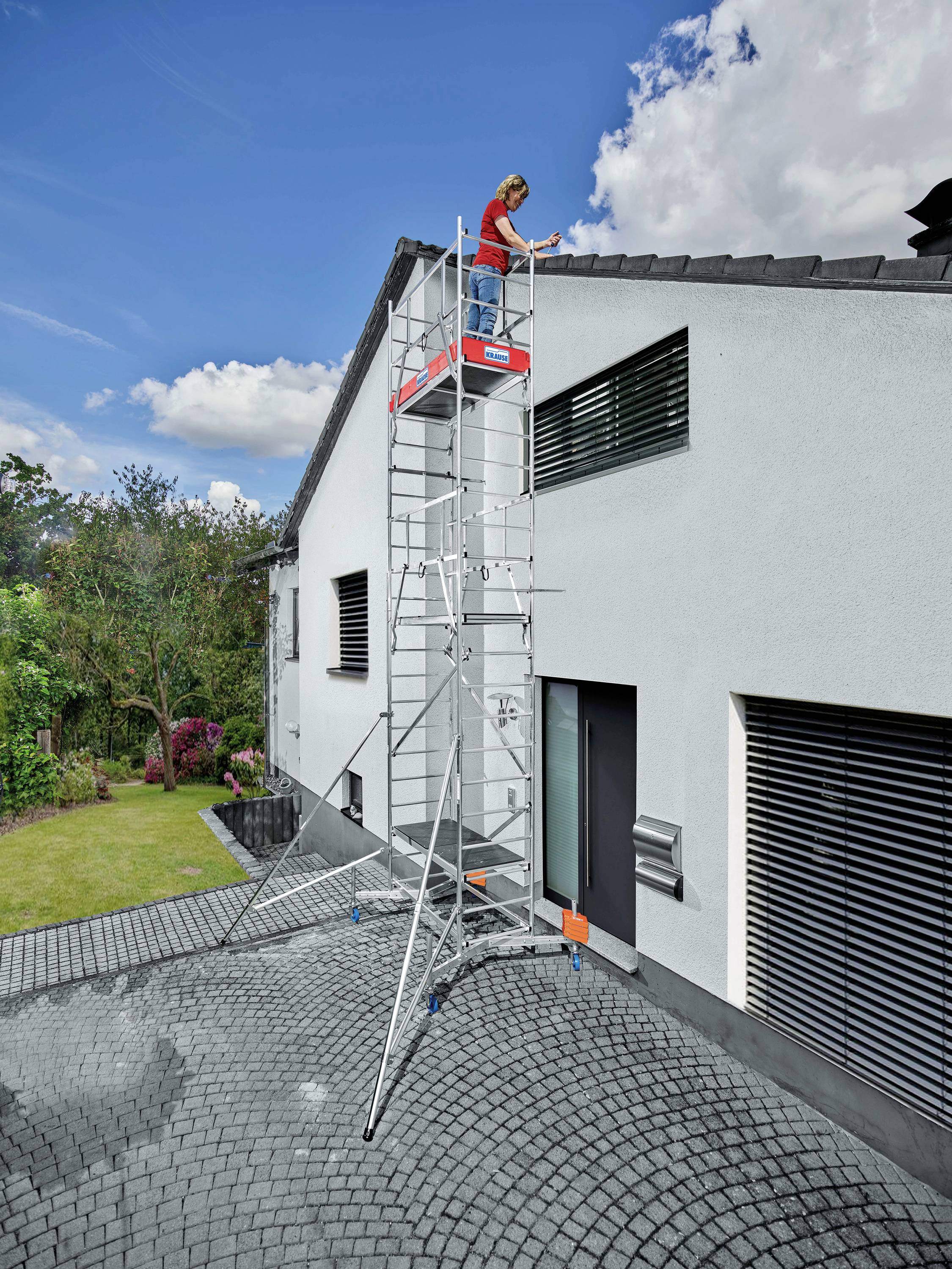 Eine Person steht auf einer hohen, stabilen Leiter an der Seitenwand eines Hauses und arbeitet am Dach. Die Umgebung ist ein Garten.